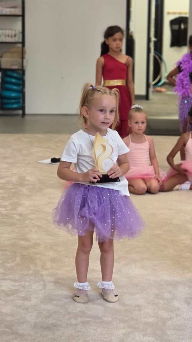 A young gymnast in a purple tutu holding up her show trophy